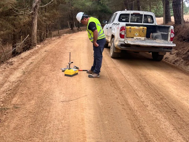Profesional con casco y chaleco reflectante realizando un ensayo de compactación con densímetro sobre un camino de tierra, junto a una camioneta
